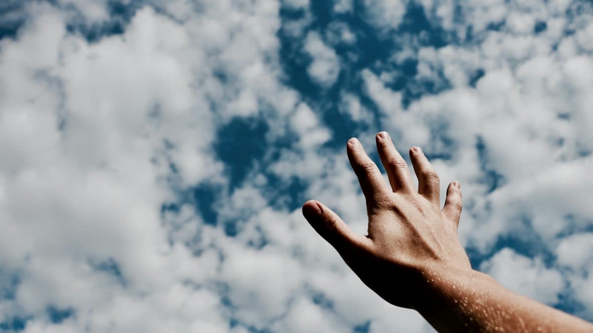 A hand reaching up toward a bright, cloud-filled sky